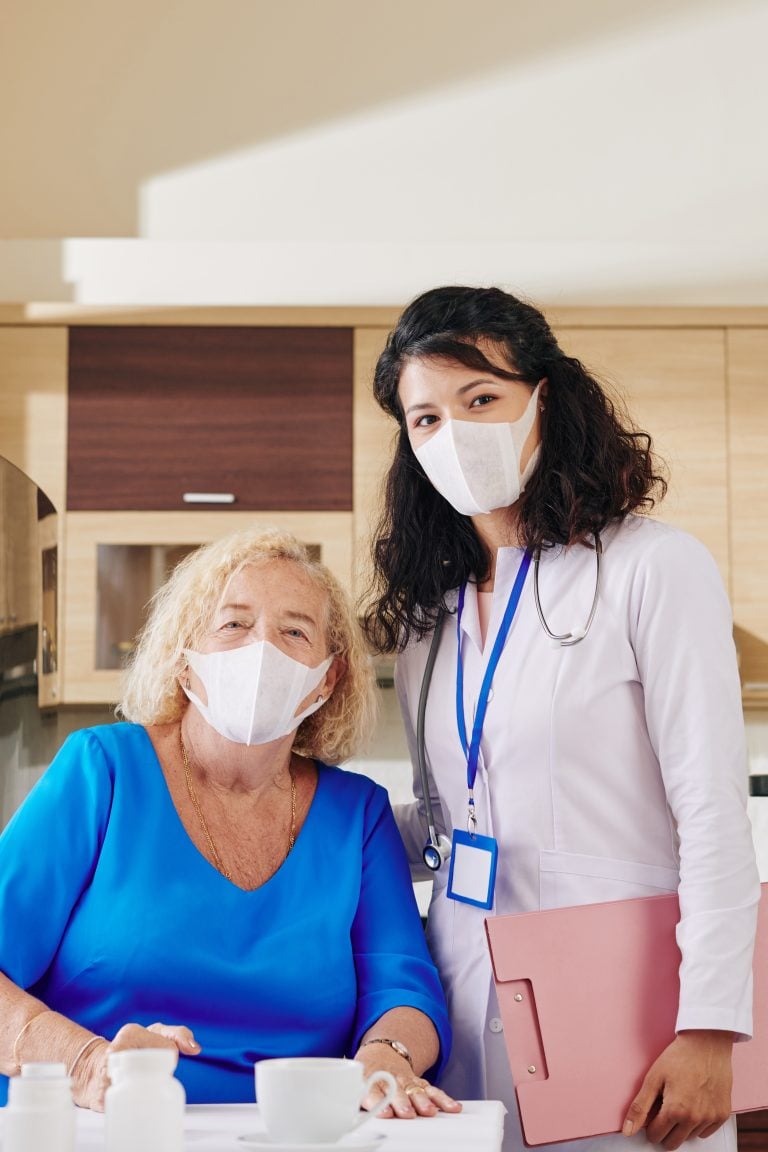 An older woman and a younger female doctor pose for the camera, both wearing masks